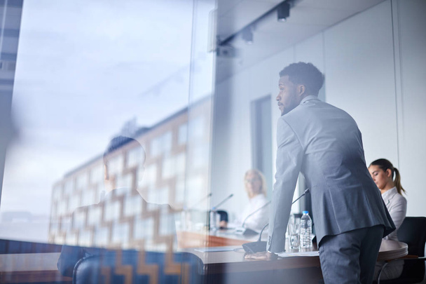 Jovem empresário afro-americano debruçando-se sobre a mesa com microfone enquanto ouve colega durante o debate
 - Foto, Imagem