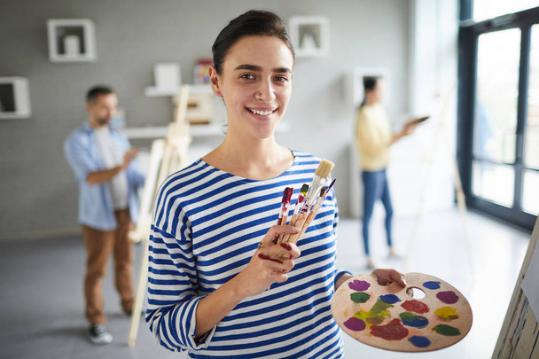 Cute girl in casualwear looking at you with smile while standing by easel and going to paint with watercolors on palette - Photo, Image