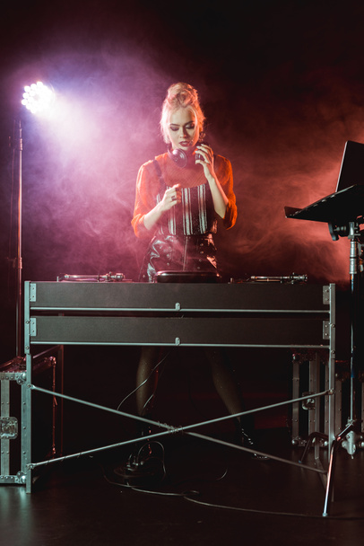 atractiva y elegante chica dj en los auriculares mirando dj mezclador en discoteca con humo
  - Foto, imagen