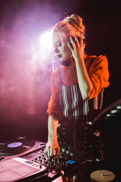 stylish dj girl in headphones standing near dj mixer in nightclub with smoke  - Photo, Image