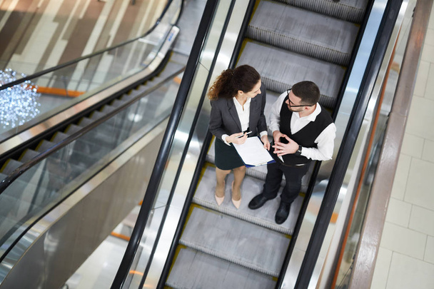Overview of two young elegant brokers standing on stairs of escalator and discussing financial document - 写真・画像