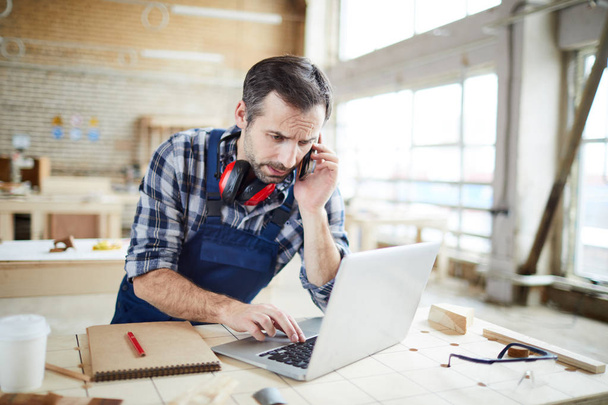 Confused frowning middle-aged bearded carpenter with ear protectors on neck standing at table and using laptop while checking information and talking by phone - Photo, Image
