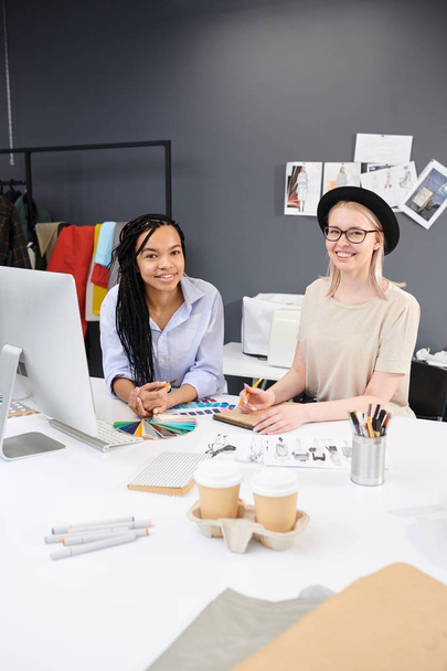 Πορτραίτο δύο χαμογελαστών σχεδιαστών μόδας που κάθονται στο τραπέζι σχεδιάζοντας σκίτσα και δουλεύοντας με πολύχρωμη παλέτα στο στούντιο - Φωτογραφία, εικόνα