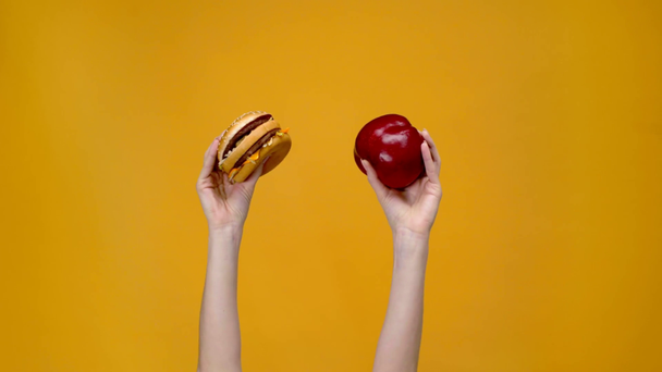 cropped view of woman presenting burger and apple isolated on yellow - Footage, Video
