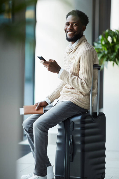 Веселый молодой путешественник в светло-бежевом свитере и серых джинсах сидит на чемодане в ожидании объявления рейса
 - Фото, изображение