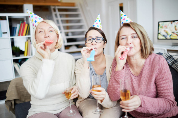 Retrato de tres mujeres felices tocando cuernos de fiesta y mirando a la cámara mientras celebran su cumpleaños en casa
 - Foto, imagen