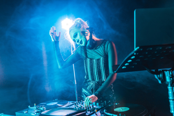 lächelndes blondes DJ-Mädchen mit Kopfhörern steht neben DJ-Mischpult in Nachtclub mit Rauch  - Foto, Bild
