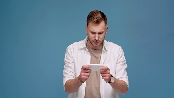 gutaussehender konzentrierter Mann, der ein Spiel auf dem Smartphone spielt, isoliert auf blauem Grund - Filmmaterial, Video