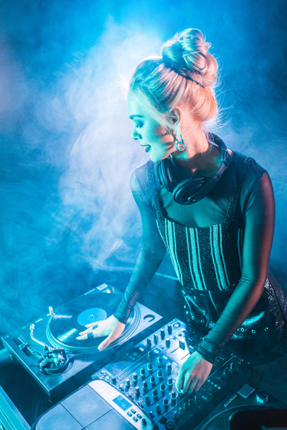happy blonde dj girl in headphones standing near dj mixer and vinyl record in nightclub with smoke  - Foto, Imagen