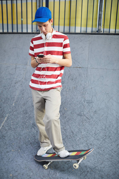 Guy in casual t-shirt, pants, baseball cap and cross-shoes messaging in smartphone during chill on skateboard - Foto, afbeelding