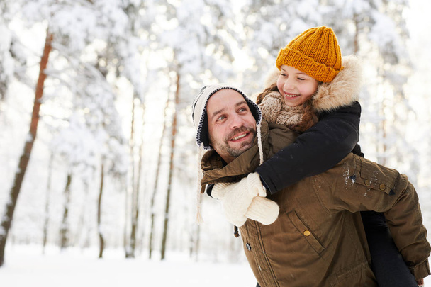 Porträt eines glücklichen kleinen Mädchens, das auf dem Rücken von Vätern reitet und dabei Spaß im Winterwald hat, Kopierraum - Foto, Bild