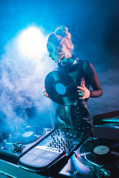 Kobieta blonde dj patrząc na sprzęt dj i przytrzymanie retro płyta winylowa w klubie nocnym z dymu  - Zdjęcie, obraz