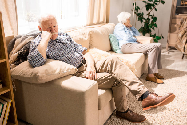 Ganzkörperporträt moderner Senioren, die auf gegenüberliegenden Seiten der Couch sitzen, Fokus auf nachdenklichen alten Mann im Vordergrund, Kopierraum - Foto, Bild