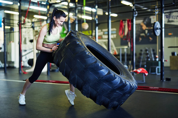 Młoda silna aktywna kobieta w odzieży sportowej podnosząca duże ciężkie opony podczas treningu w centrum rekreacyjnym - Zdjęcie, obraz