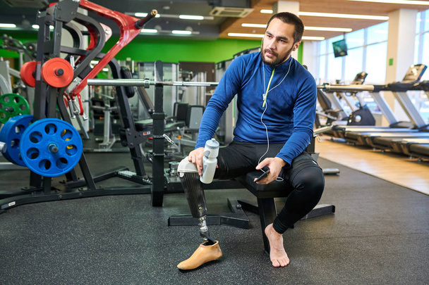 Νέος όμορφος αθλητής με ακρωτηριασμένο πόδι κάθεται στο γυμναστήριο με ένα μπουκάλι νερό και ακούγοντας μουσική στο smartphone του - Φωτογραφία, εικόνα