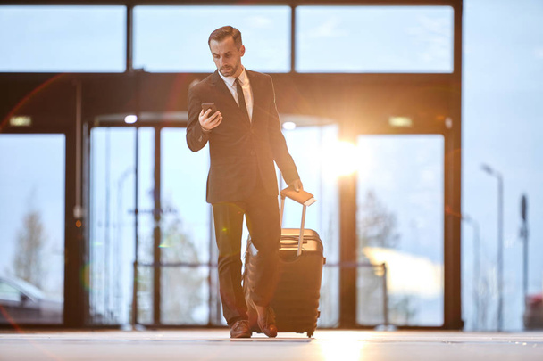 junger eleganter Geschäftsmann zieht Koffer, während er vor der Abreise im Smartphone blättert - Foto, Bild