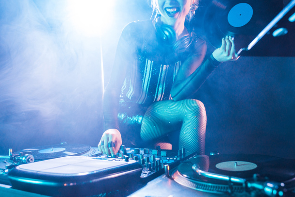 cropped view of cheerful dj woman holding retro vinyl record and standing near dj equipment in nightclub with smoke  - Foto, immagini