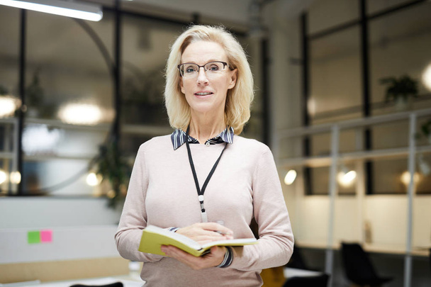Sebevědomá blondýnka v brýlích, která stojí v moderní kanceláři a drží otevřený deník při pohledu na fotoaparát - Fotografie, Obrázek
