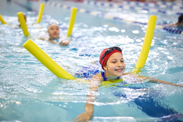 Klein meisje zwemmen met noodle in het binnenzwembad en genieten van het proces tijdens het zwemmen les - Foto, afbeelding