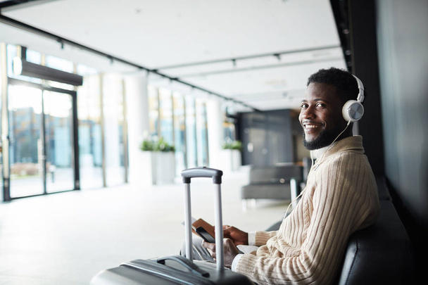 Sonriente hombre de negocios afroamericano positivo en ropa casual sentado en el salón del aeropuerto y disfrutando de la música en los auriculares
 - Foto, imagen