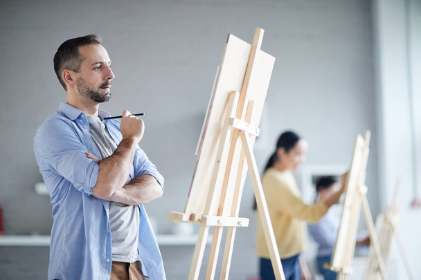 Молодий чоловік зосередився на створенні ескізу на мольберті перед живописом під час уроку в художній студії
 - Фото, зображення