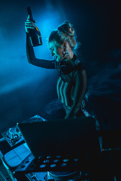 žena veselá blondýnka dj sluchátka drží láhev a usmívá se poblíž dj vybavení v nočním klubu s kouřem - Fotografie, Obrázek