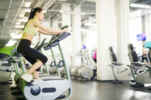 Young active woman in sportswear sitting on one of sports bicycles and exercising in fitness center - Photo, Image