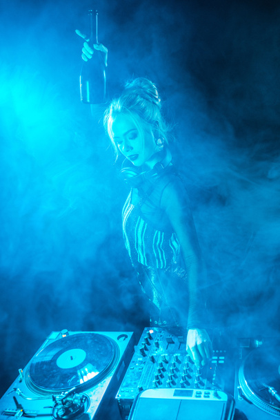 blonde dj woman in headphones holding  bottle near dj equipment in nightclub with smoke - Foto, Imagem