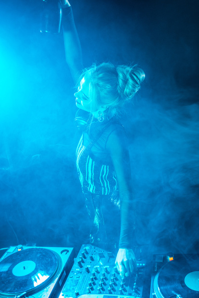 щаслива блондинка джи в навушниках тримає пляшку біля обладнання джи в нічному клубі з димом
 - Фото, зображення