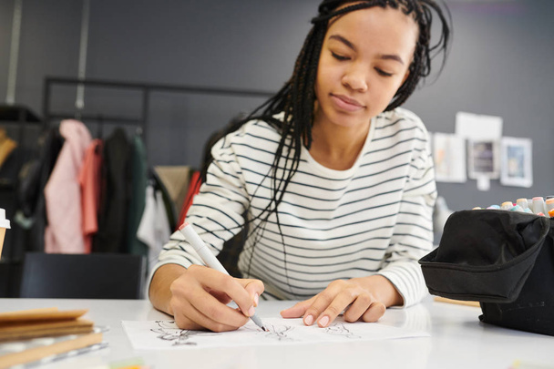 Genç bayan tasarımcı masada oturuyor ve tasarım stüdyosunda kağıt üzerine kıyafet çizimleri yapıyor. - Fotoğraf, Görsel