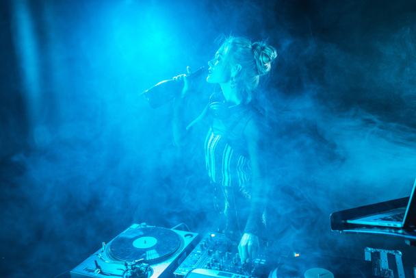 happy blonde dj woman in headphones drinking wine from  bottle near dj equipment in nightclub with smoke - Fotoğraf, Görsel