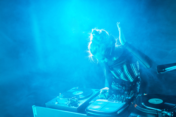 blonde dj girl listening music in headphones while using dj equipment in nightclub with smoke  - Foto, afbeelding