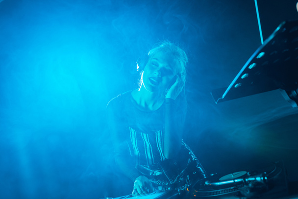 glückliches blondes DJ-Mädchen, das Musik in Kopfhörern mit geschlossenen Augen in einem Nachtclub mit Rauch hört  - Foto, Bild