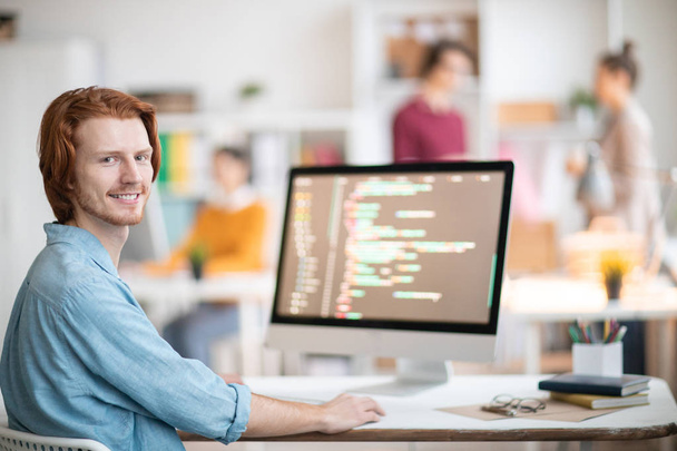 Joven diseñador o programador exitoso mirándote mientras estás sentado junto al escritorio y decodificando datos en línea
 - Foto, imagen
