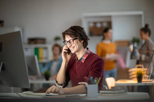 Lächelnder junger Mann in Freizeitkleidung sitzt vor dem Computerbildschirm und berät sich mit seinem Kollegen per Smartphone - Foto, Bild