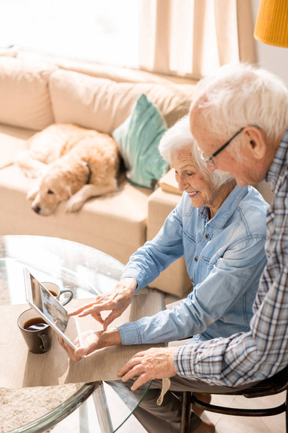 Portret o wysokim kącie nowoczesnej pary seniorów przy użyciu cyfrowego tabletu siedzącego przy stole w domu z psem w tle - Zdjęcie, obraz