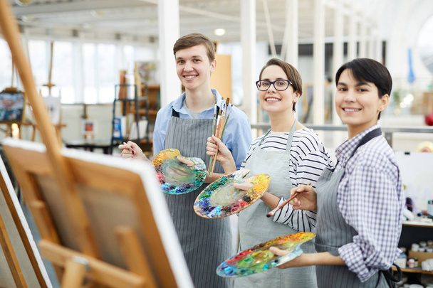 Taillenhoch Porträt von drei fröhlichen Kunststudenten, die mit Staffeleien und Paletten im Atelier vor der Kamera posieren - Foto, Bild