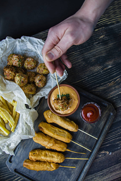 The male hand inserts cabbage croquet into the sauce. Fried french fries, dog corn and cabbage croquettes and potatoes with sauce, ketchup decorated with fresh vegetables and greens in the background. - Photo, image