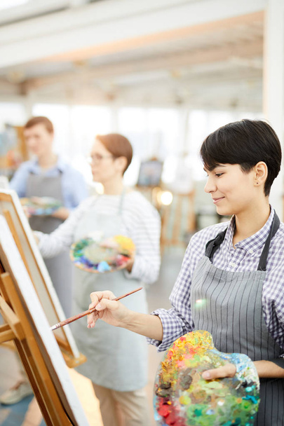 Portret z boku młodej kobiecej artysty trzymającej paletę i obraz malarstwa na sztalidie korzystających z pracy w Studio sztuki - Zdjęcie, obraz