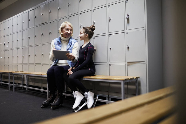 Ganzkörperporträt der kleinen Eiskunstläuferin mit digitalem Tablet in der Umkleidekabine mit Mama oder Trainer nach dem Training, Kopierraum - Foto, Bild