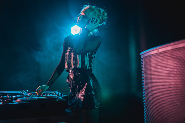 happy dj woman with blonde hair listening music and touching headphones in nightclub with smoke - Photo, Image
