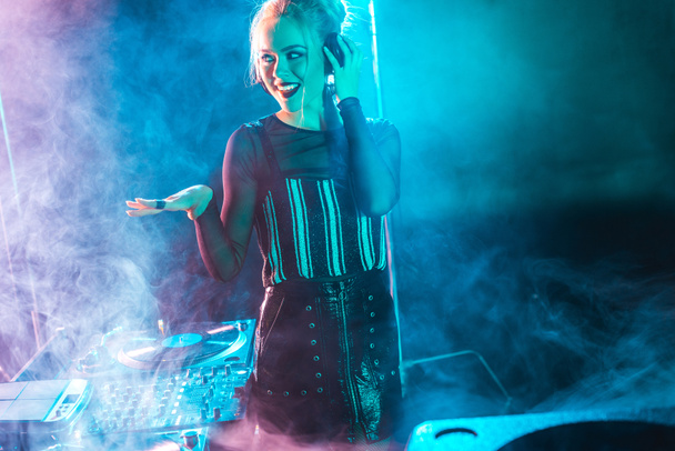 happy dj woman with blonde hair listening music and touching headphones in nightclub with smoke - Foto, Bild
