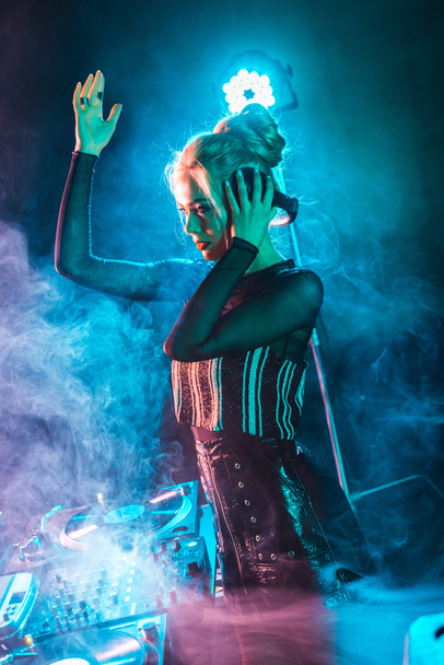 hermosa mujer dj con cabello rubio escuchando música y sosteniendo auriculares y gestos en discoteca con humo
 - Foto, Imagen