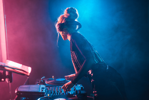 dj woman with blonde hair using dj mixer and touching vinyl record in nightclub with smoke - Foto, afbeelding