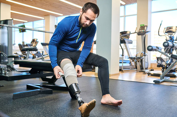 Guapo atleta barbudo sentado y vendándose la pierna antes de entrenar en el gimnasio
 - Foto, imagen