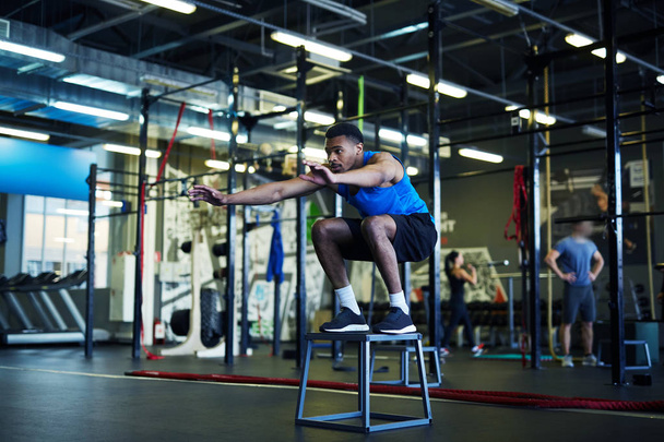 Jeune sportif afro-américain debout sur une chaise de saut avec les genoux pliés et les bras tendus
 - Photo, image