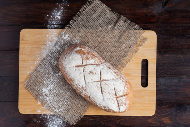 frisches braunes Brot auf einem Eichenbrett mit verstreutem Mehl auf einem hölzernen Hintergrund mit einem Stück Leinenstoff - Foto, Bild
