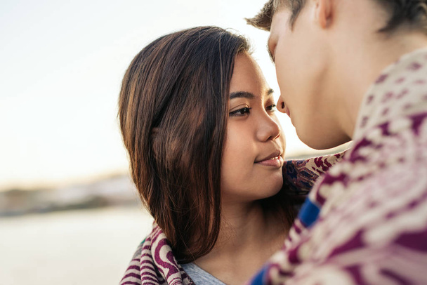 Affectionate young couple about to kiss while wrapped in a blanket together by a harbor on an autumn afternoon - 写真・画像