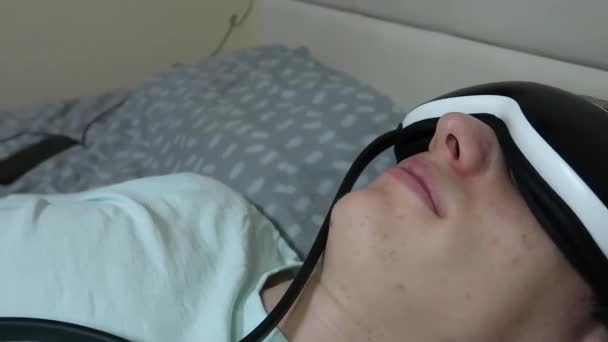 joven atractivo hombre utiliza dispositivo masajeador de ojos, acostado en la cama, sosteniendo un control remoto, aliviar la fatiga, relajación, descanso después de trabajar en un ordenador portátil, primer plano - Metraje, vídeo