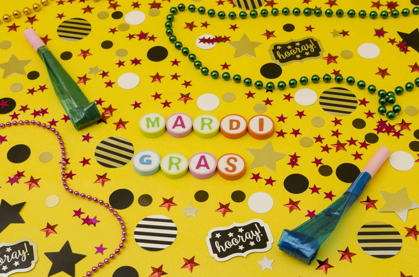 Top view decoration for party. Mardi Gras made from colorful letters, plastic necklaces, party blowers and circles and stars confetti on yellow background - Photo, Image